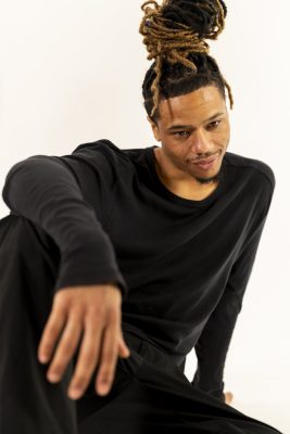 Danseur aux dreadlocks assis au sol, la main tendue vers l'objectif